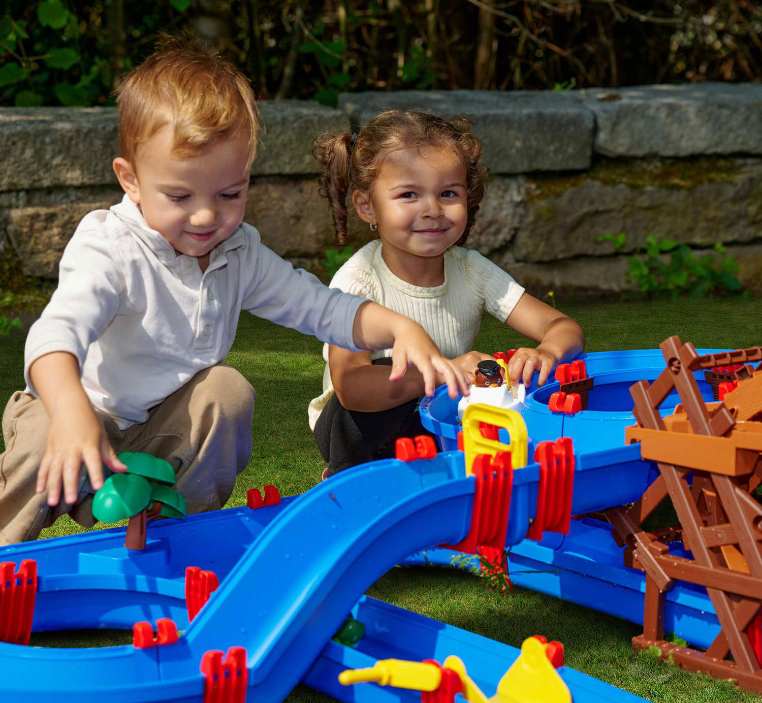 Zwei kleine Kinder spielen mit einer blauen Wasserbahn im Garten.