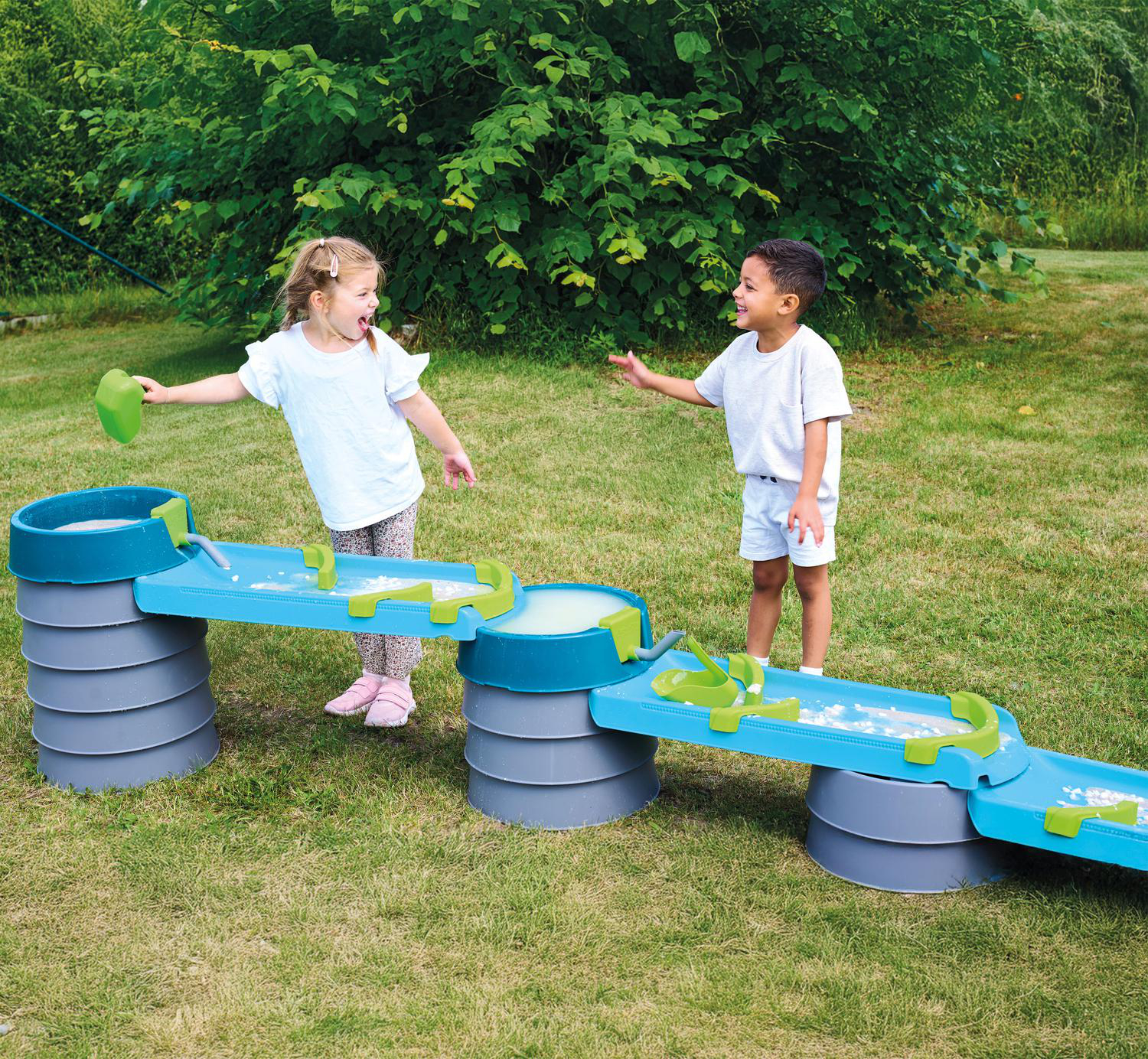 Zwei Kinder spielen mit einem blauen und grünen Wasserspieltisch im Garten.