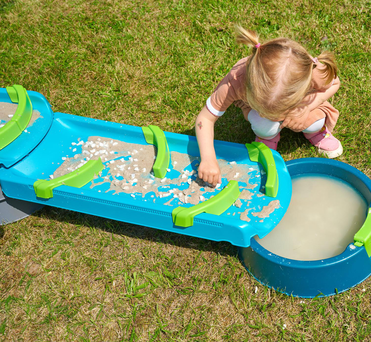 Ein kleines Mädchen spielt mit einem blau-grünen Wassertisch voller Sand und Steine.