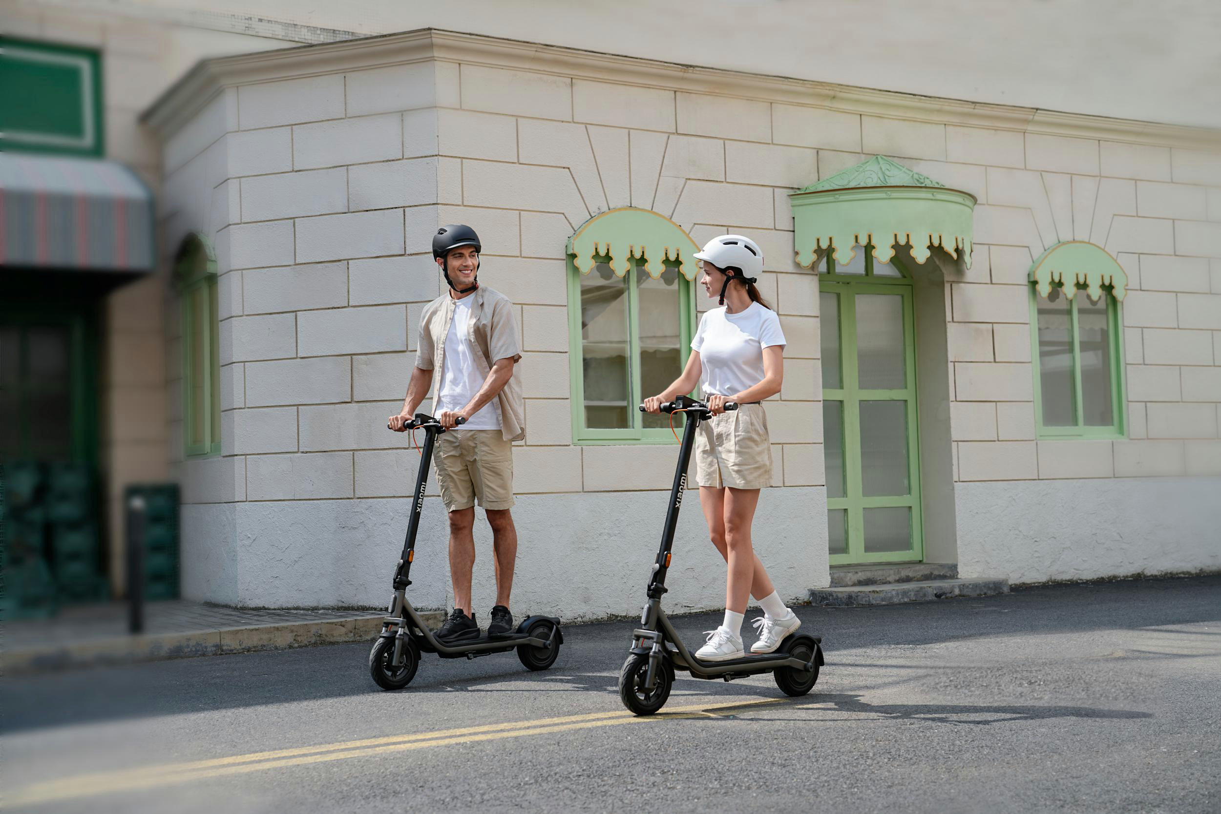 Ein Mann und eine Frau fahren E-Scooter vor einem Gebäude mit grünen Fenstern.