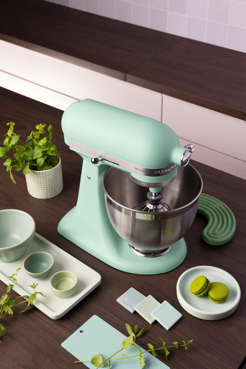 Hellgrüner Küchenmixer mit Edelstahlschüssel, daneben Schalen, Macarons und Pflanzen.