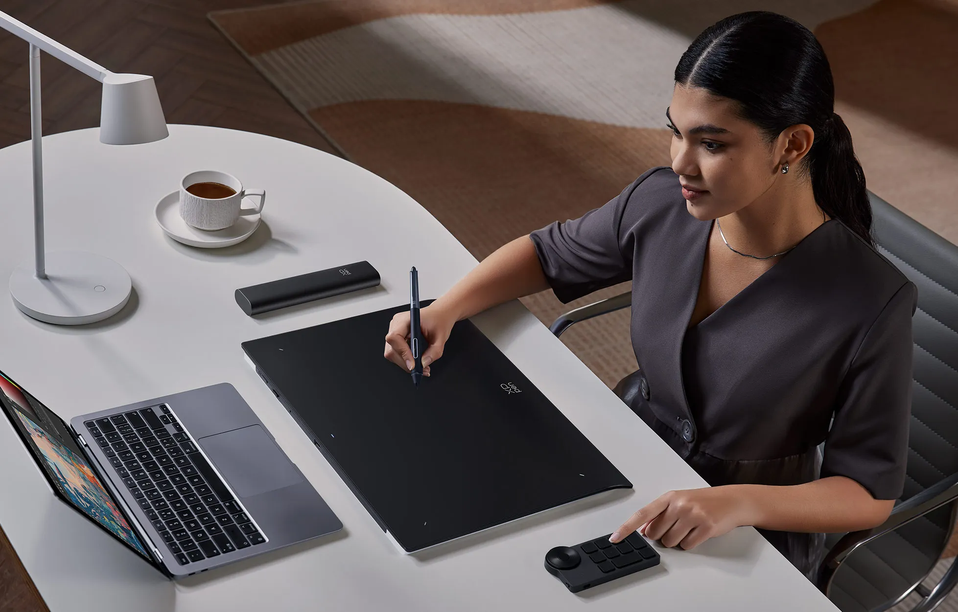 Donna usa tavoletta grafica con penna, laptop e tazza di caffè su scrivania.