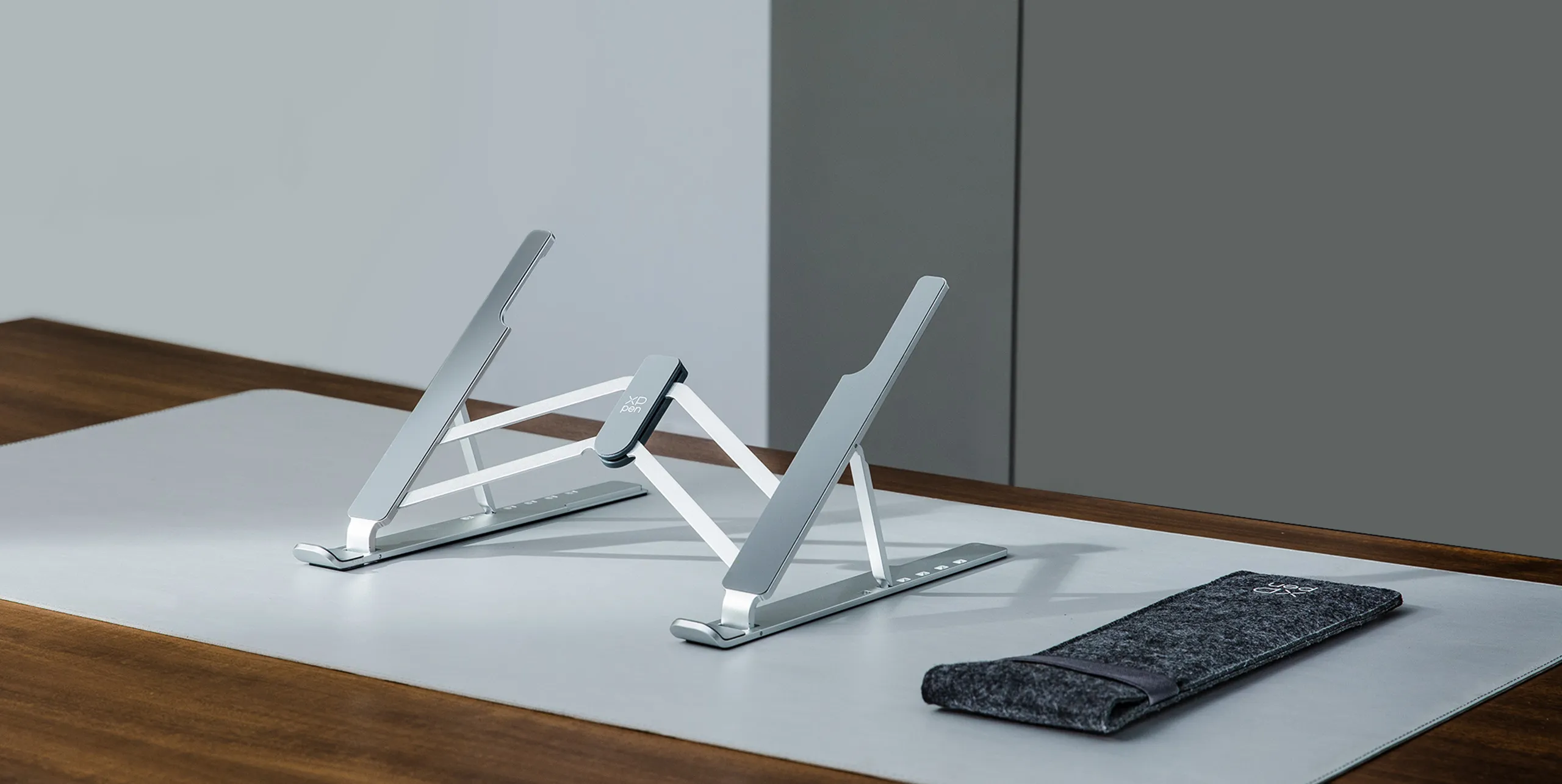 Due supporti per laptop argentati su un tappetino grigio accanto a una custodia in feltro.