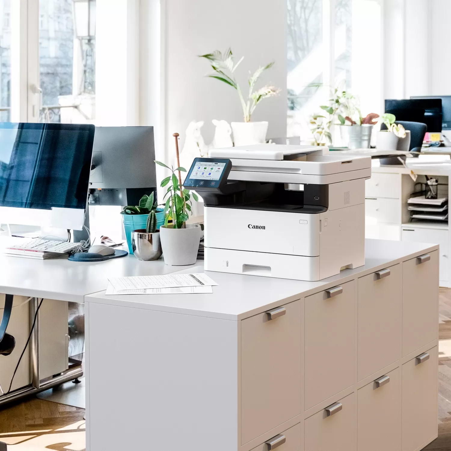 Ein weißer Canon Drucker steht auf einem Schreibtisch mit Computern und Pflanzen.