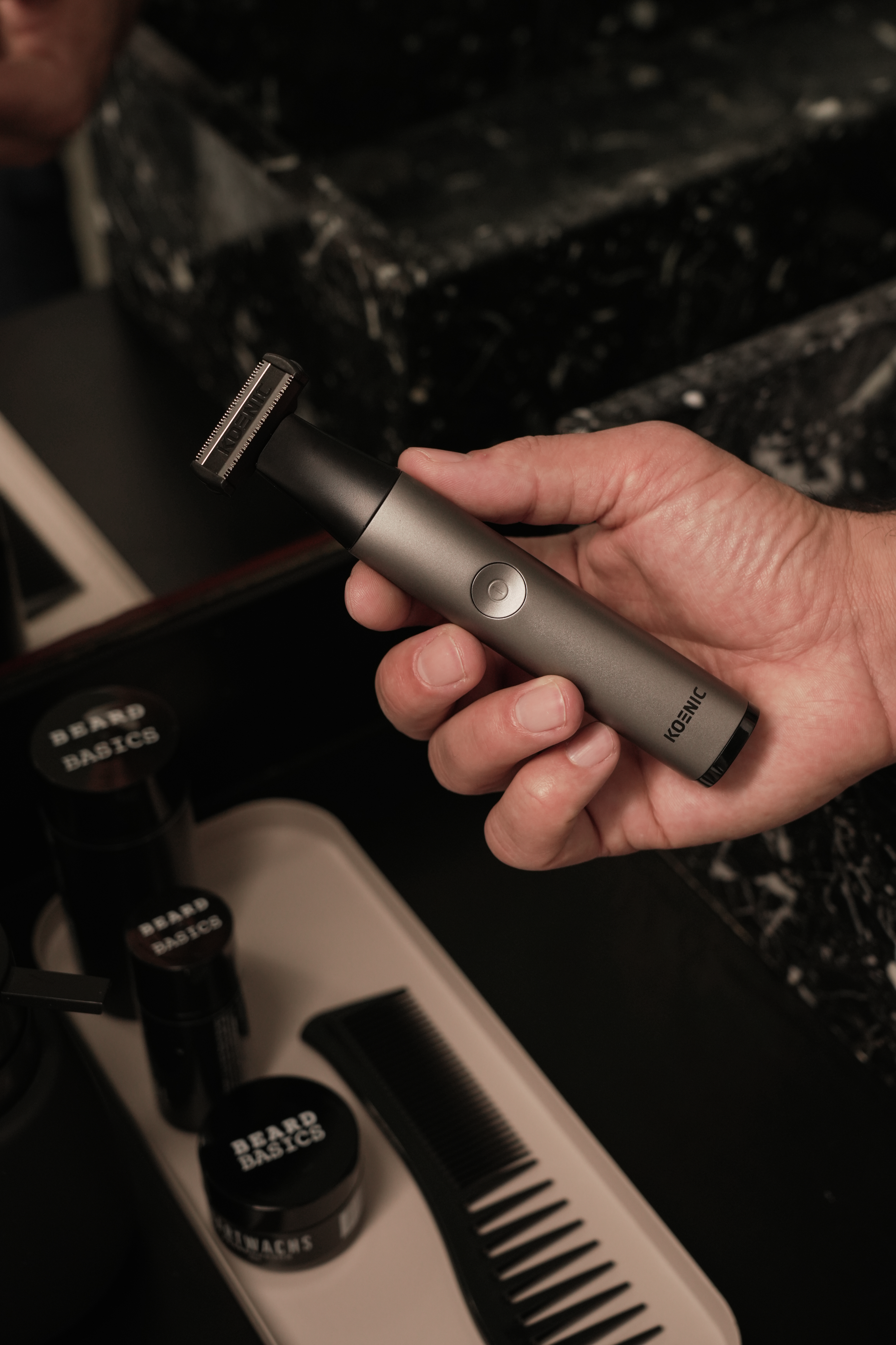 Dłoń trzyma szary trymer nad produktami do brody i grzebieniem.