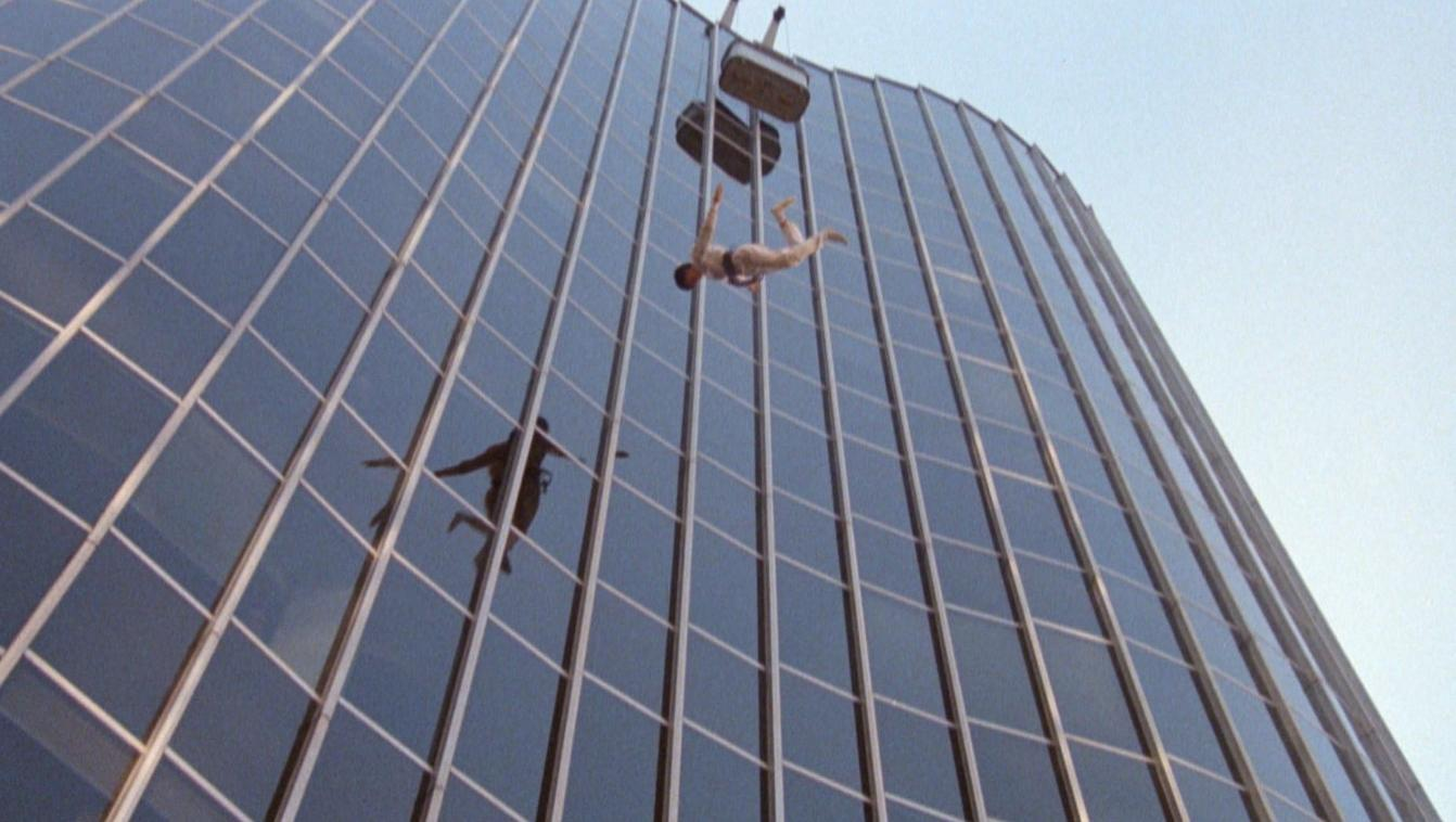 Zwei Personen fallen vor einem Glasgebäude, eine hängt an einem Kasten.