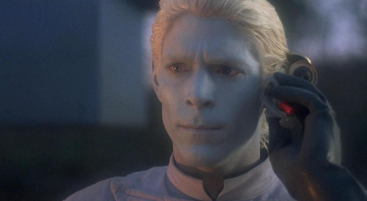 Ein Mann mit blauer Haut und blonden Haaren hält ein Gerät ans Ohr.