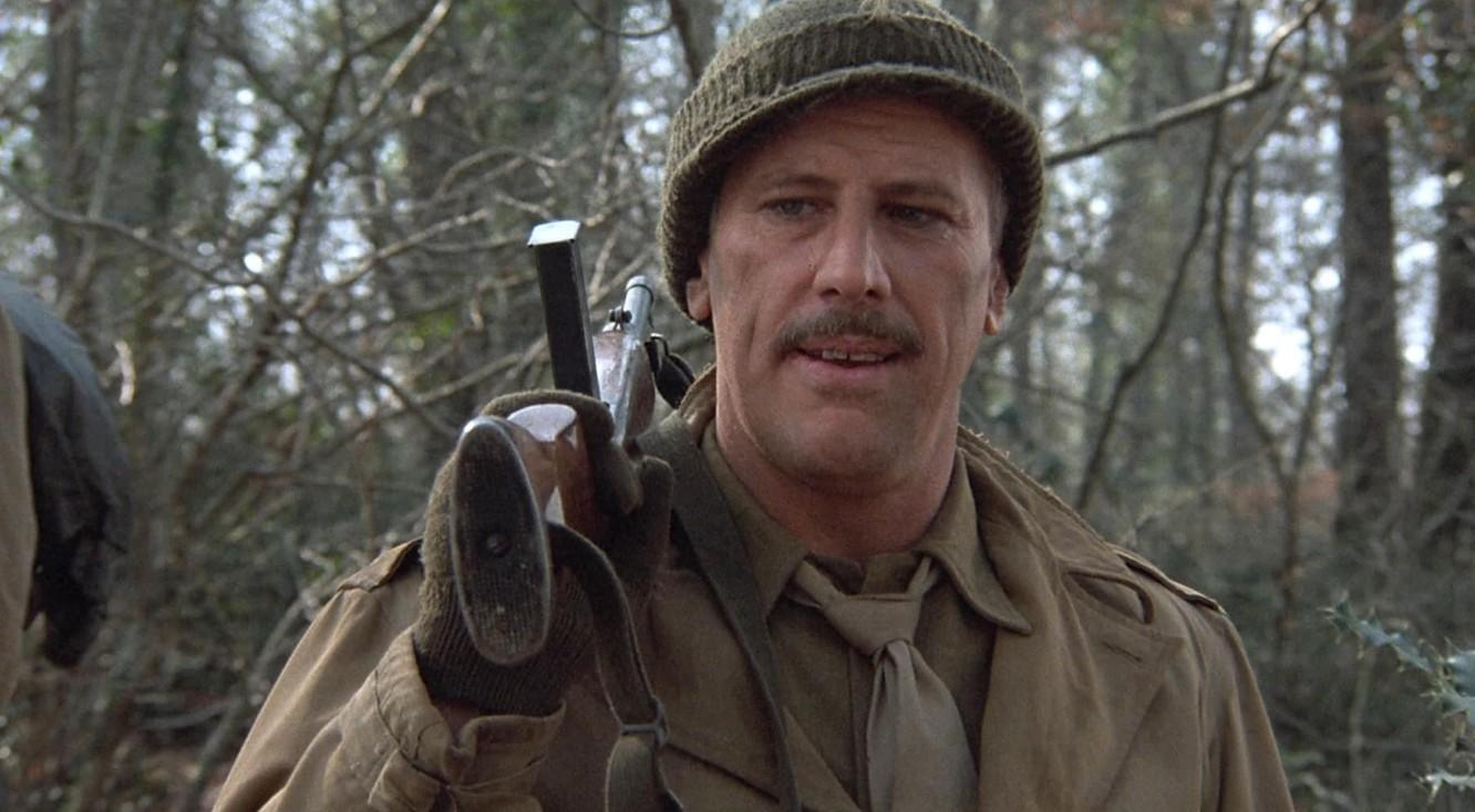 Mann in Uniform mit Gewehr im Wald. Er trägt eine Mütze und ein Band.