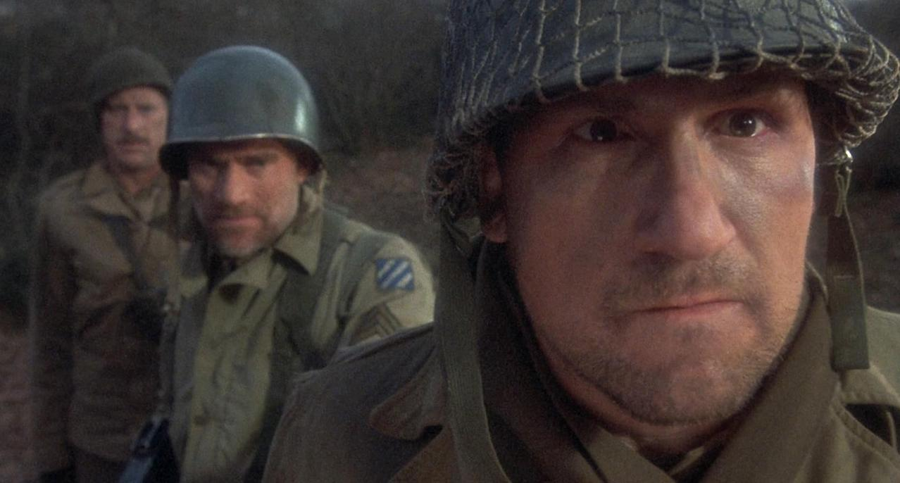 Drei Soldaten in Uniformen, einer im Vordergrund mit Helm, die anderen unscharf im Hintergrund.