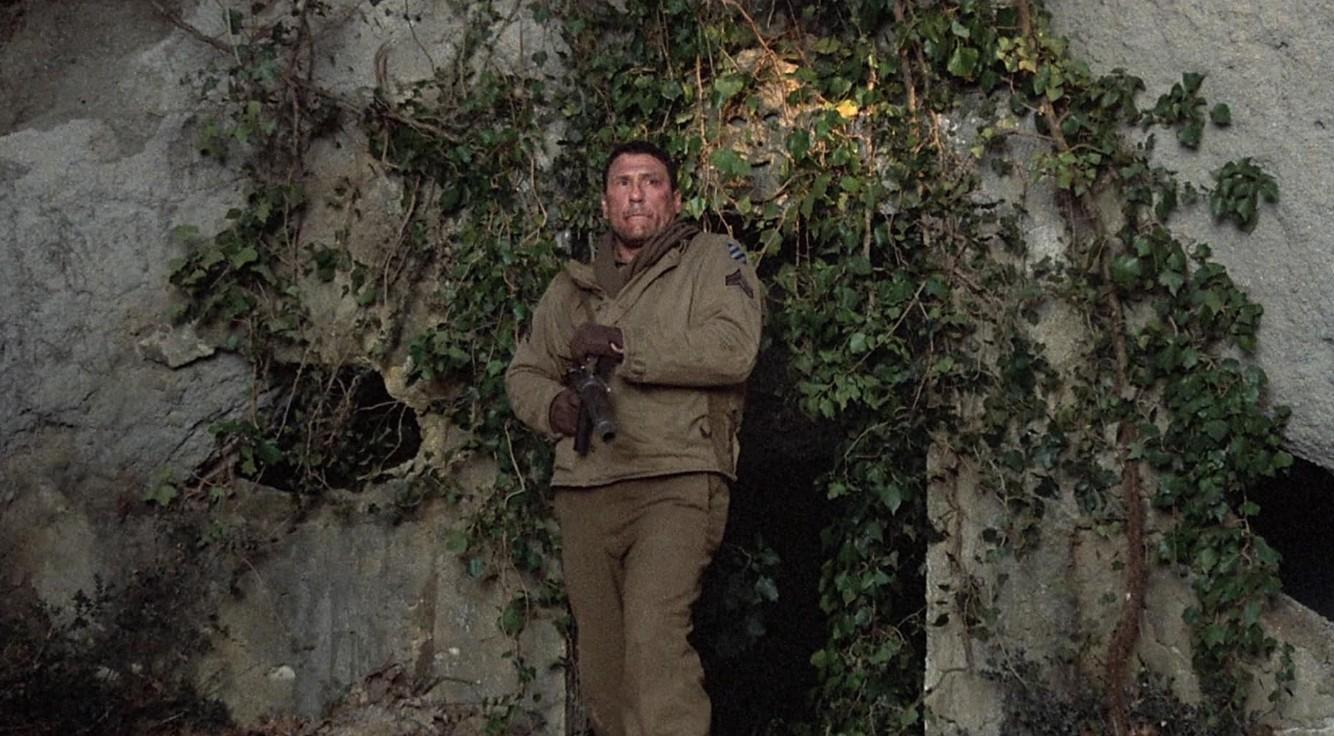 Ein Mann in Uniform mit Waffe vor einer bemoosten Felswand.