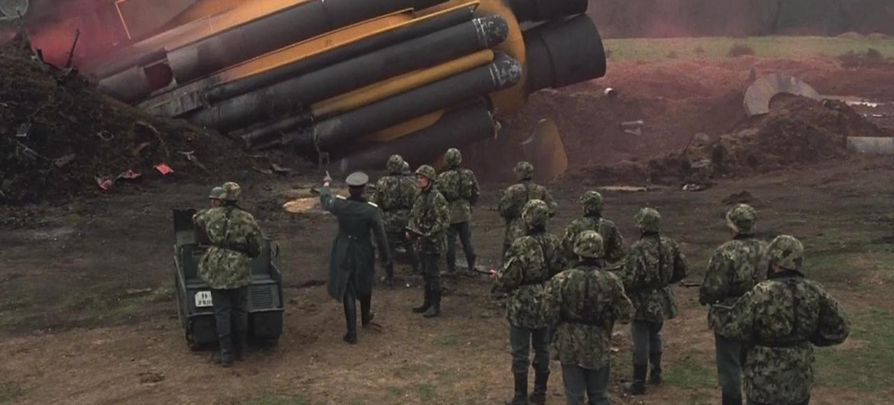 Soldaten in Tarnung stehen vor abgestürztem Flugzeugwrack mit gelben Rohren.