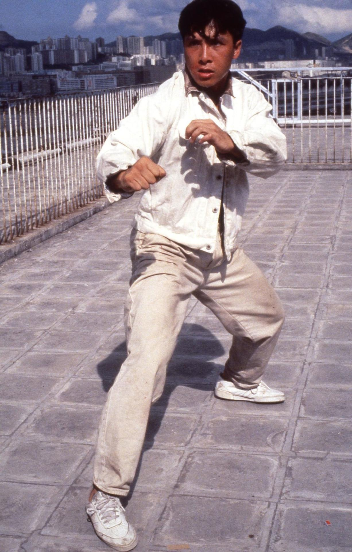 Ein Mann in weiem Hemd und heller Hose kämpft auf einem Gehweg mit Stadtblick.