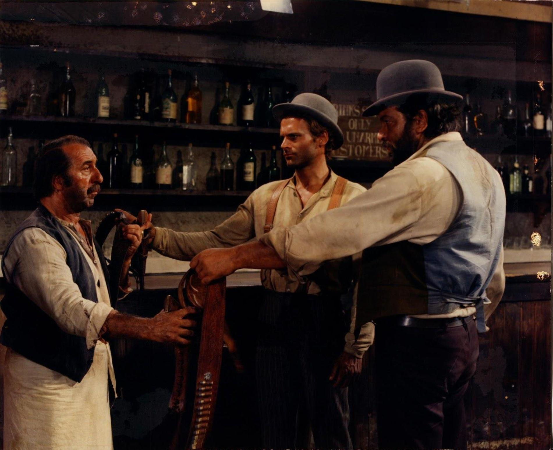 Drei Männer in einem Saloon mit Regalen voller Flaschen im Hintergrund.