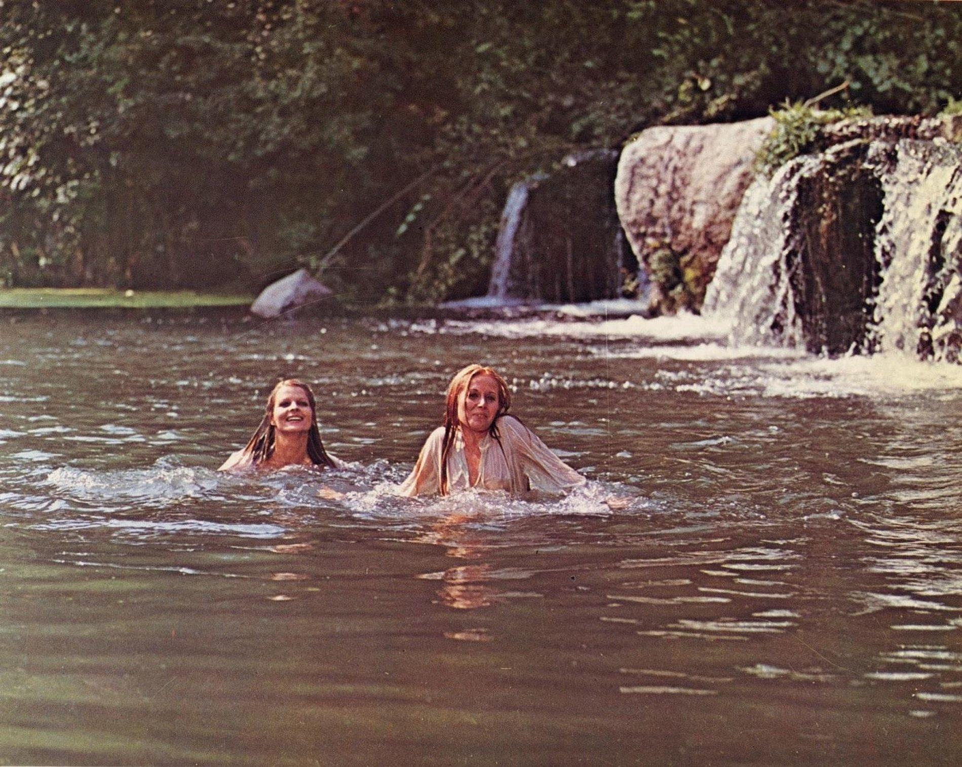 Zwei Frauen schwimmen in einem Fluss vor einem kleinen Wasserfall und Bäumen.