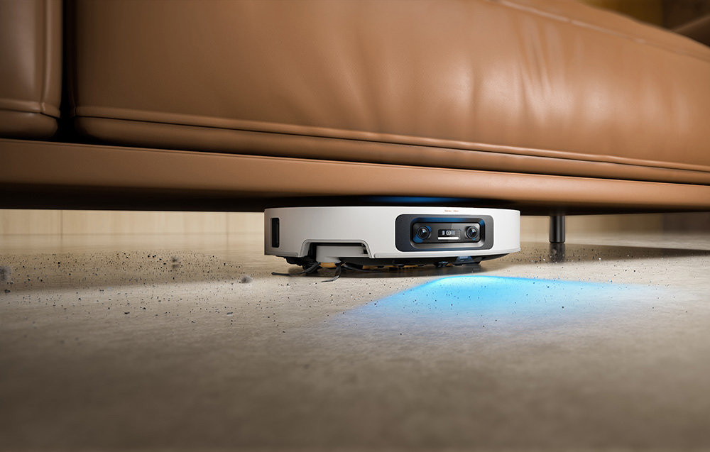 Biały robot sprzątający czyści pod brązową sofą na betonowej podłodze.