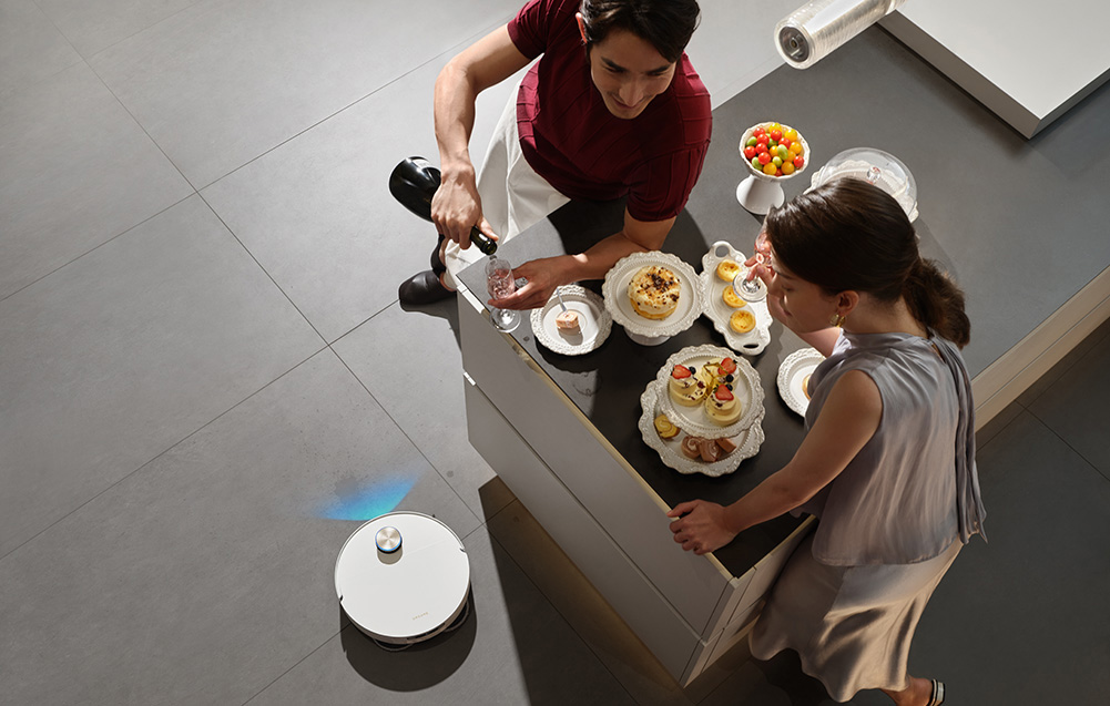 Mężczyzna nalewa szampana, podczas gdy robot odkurzający czyści podłogę.