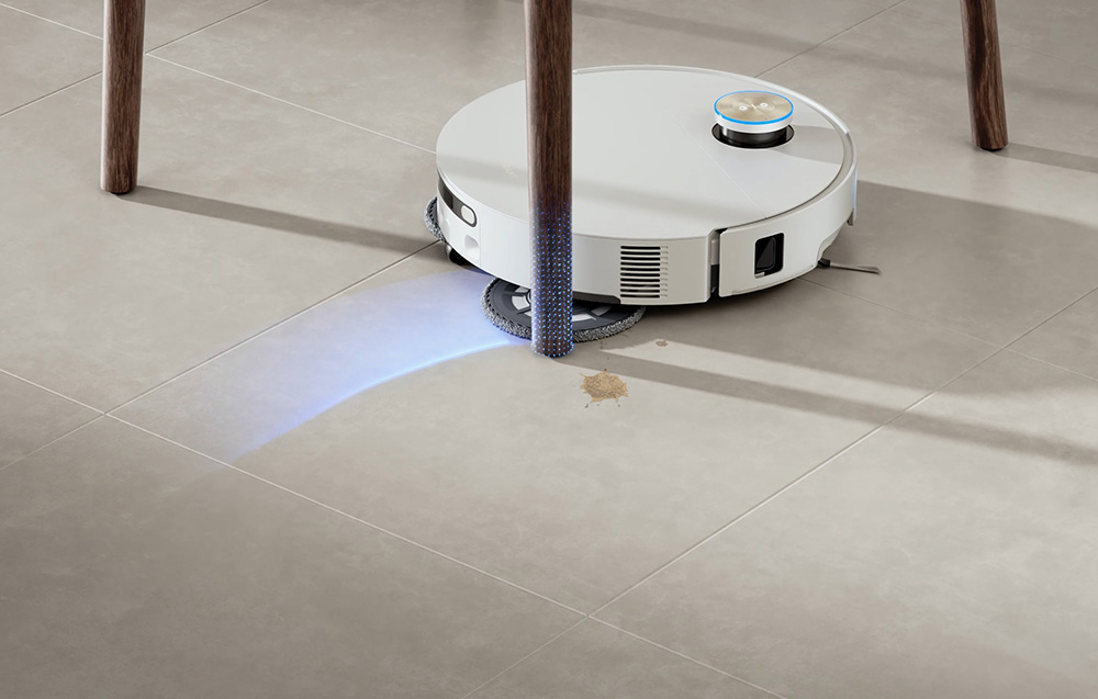 Biały robot sprzątający czyści plamę na jasnoszarej podłodze z płytek.