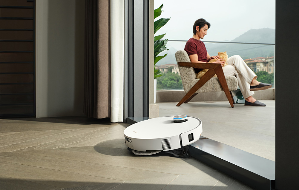 Biały robot sprzątający czyści podłogę, podczas gdy mężczyzna trzyma kota na balkonie.