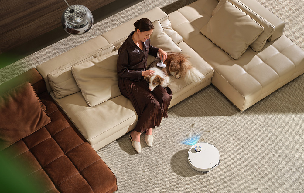 Kobieta szczotkuje psa na kanapie, robot sprzątający czyści.