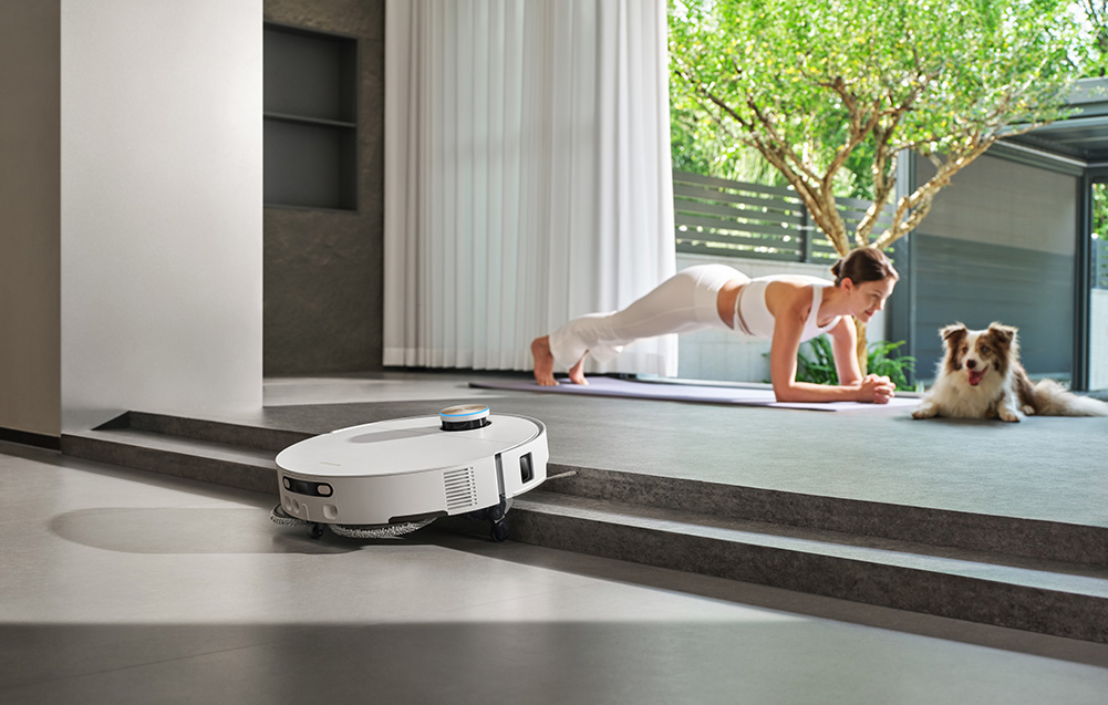 Robot sprzątający czyści podłogę, podczas gdy kobieta wykonuje deskę, a pies obserwuje.