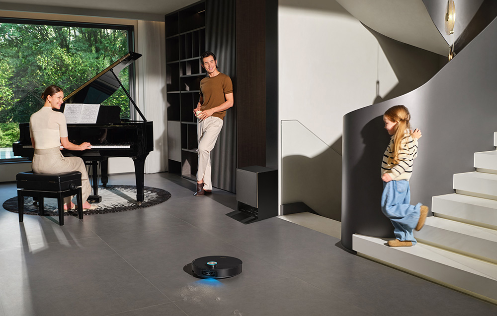 Odkurzacz automatyczny czyści kurz, kobieta gra na pianinie, mężczyzna patrzy.