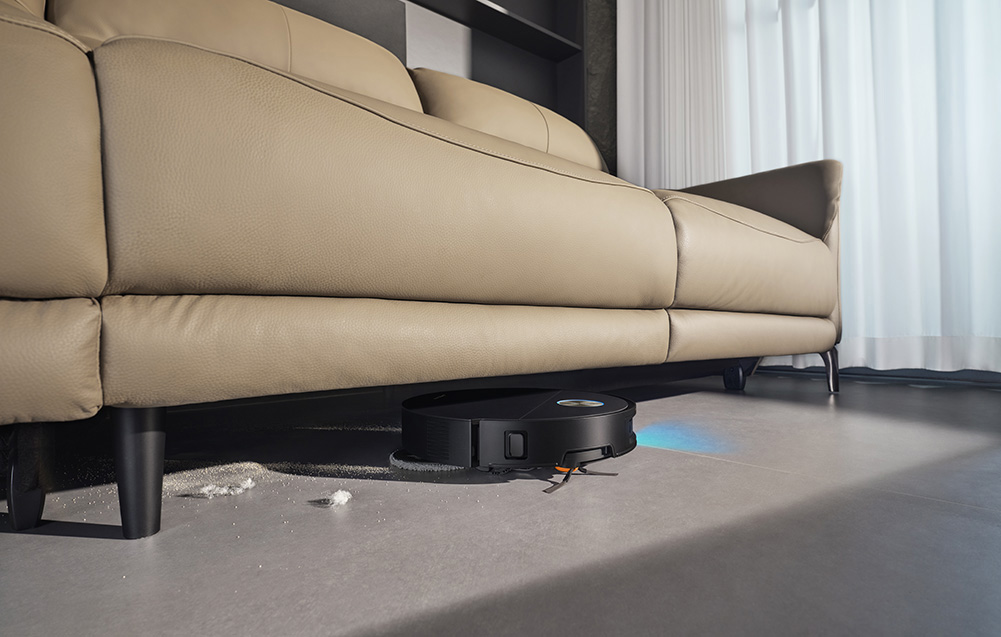 Czarny robot sprzątający czyści pod beżową sofą.
