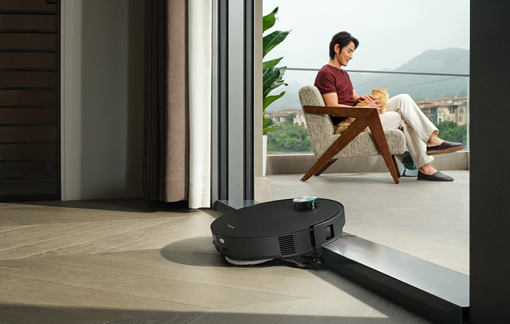 Mężczyzna z kotem siedzi na krześle, robot sprzątający czyści podłogę.