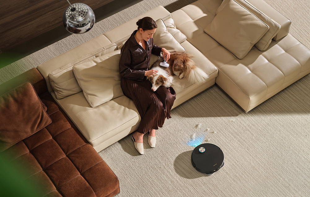 Kobieta szczotkuje psa na beżowej kanapie, podczas gdy robot odkurzający sprząta.