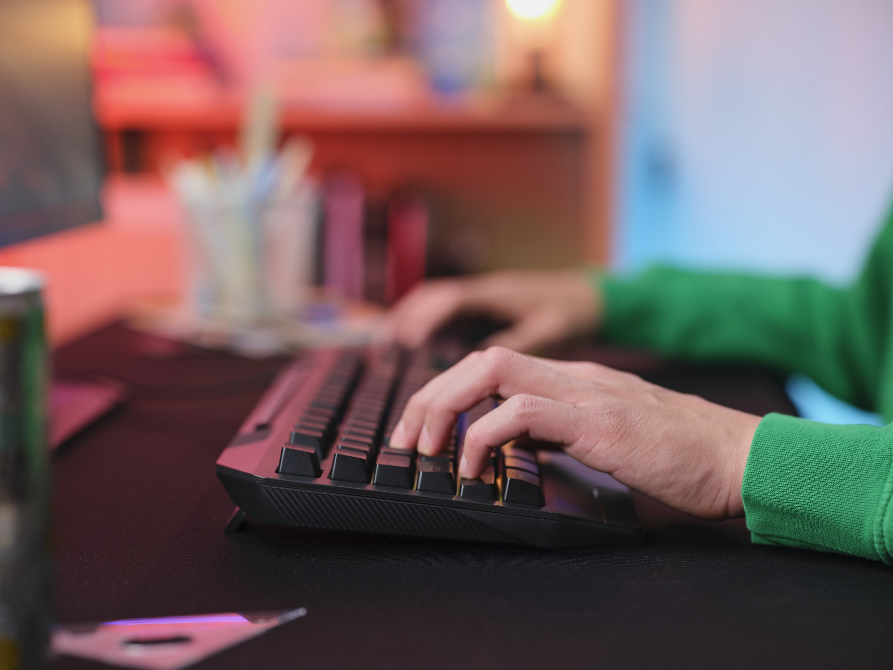 Dłonie piszą na czarnej klawiaturze, osoba w zielonej bluzie. Biurko w kolorowym świetle.