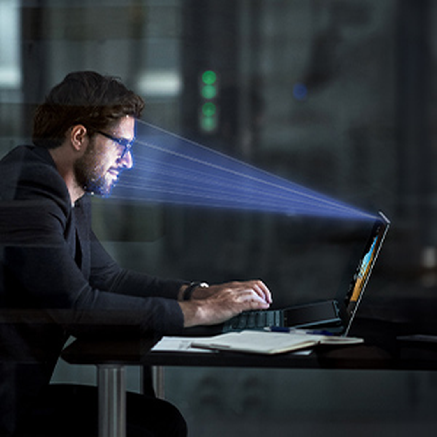 Man in glasses works on laptop, blue light beams scan his face.