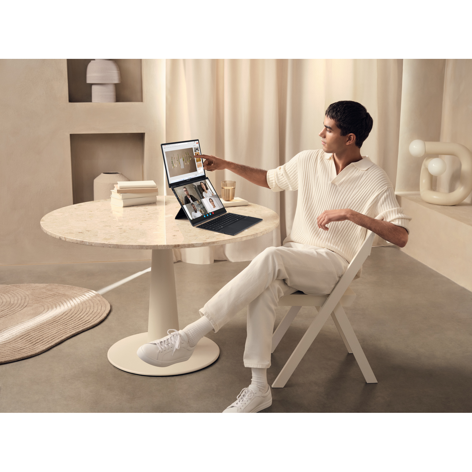 Man in white clothing sits at a table, using a laptop with three people on a video call.