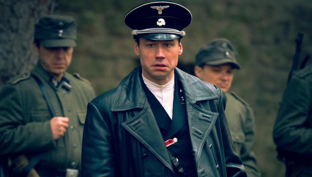 Ein Mann in SS-Uniform und schwarzem Mantel steht im Vordergrund. Andere Männer in Uniform sind im Hintergrund.