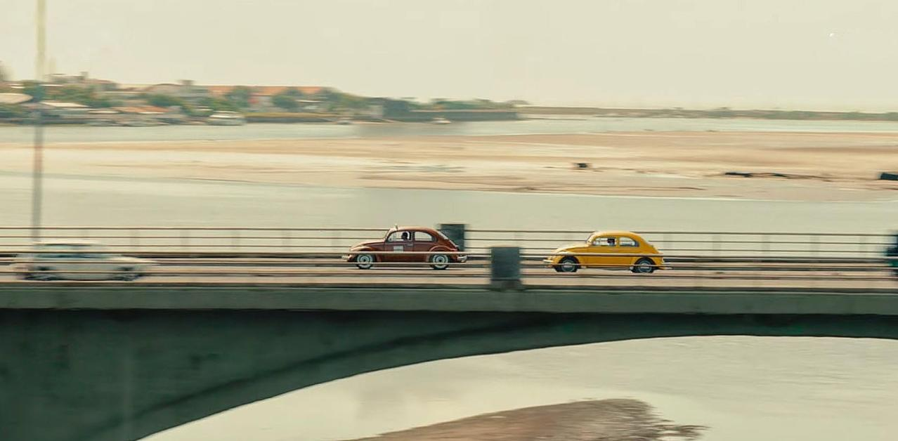 Zwei Oldtimer, braun und gelb, fahren auf einer Brücke über Wasser.