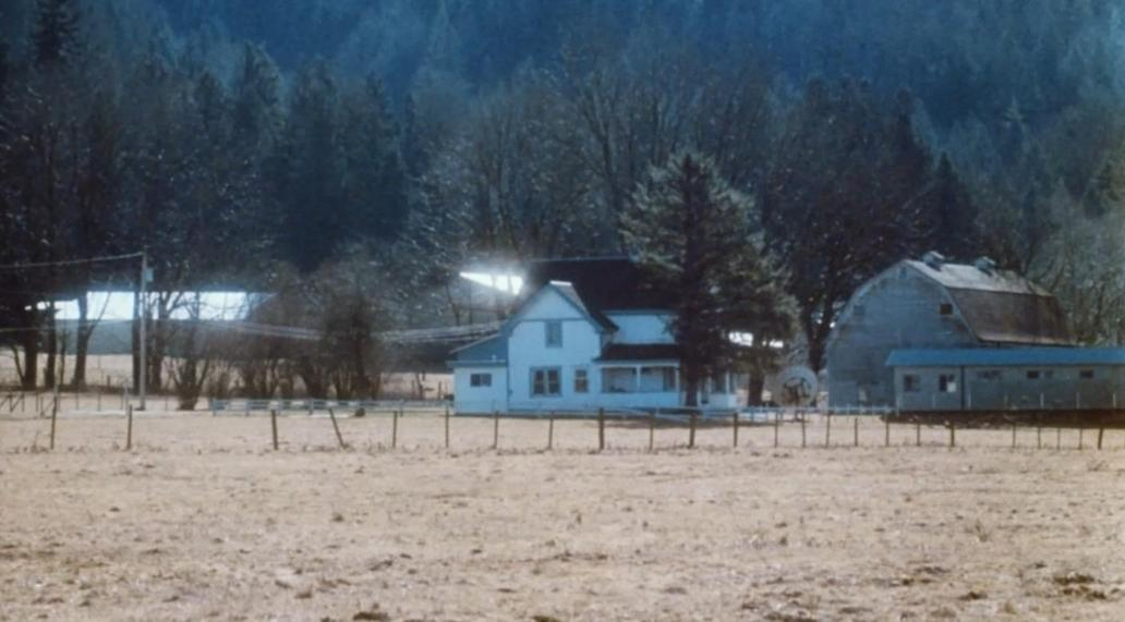Ein weißes Haus und eine Scheune stehen auf einem Feld vor einem dunklen Wald.
