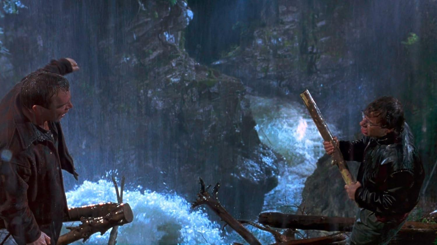 Zwei Männer stehen im Regen an einem reißenden Fluss, einer mit einem Ast.