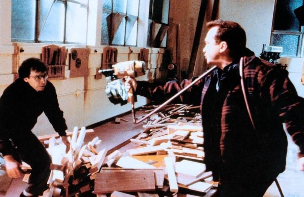 Ein Mann mit Brille weicht vor einem Mann mit einem Hammer im Werkstatt.