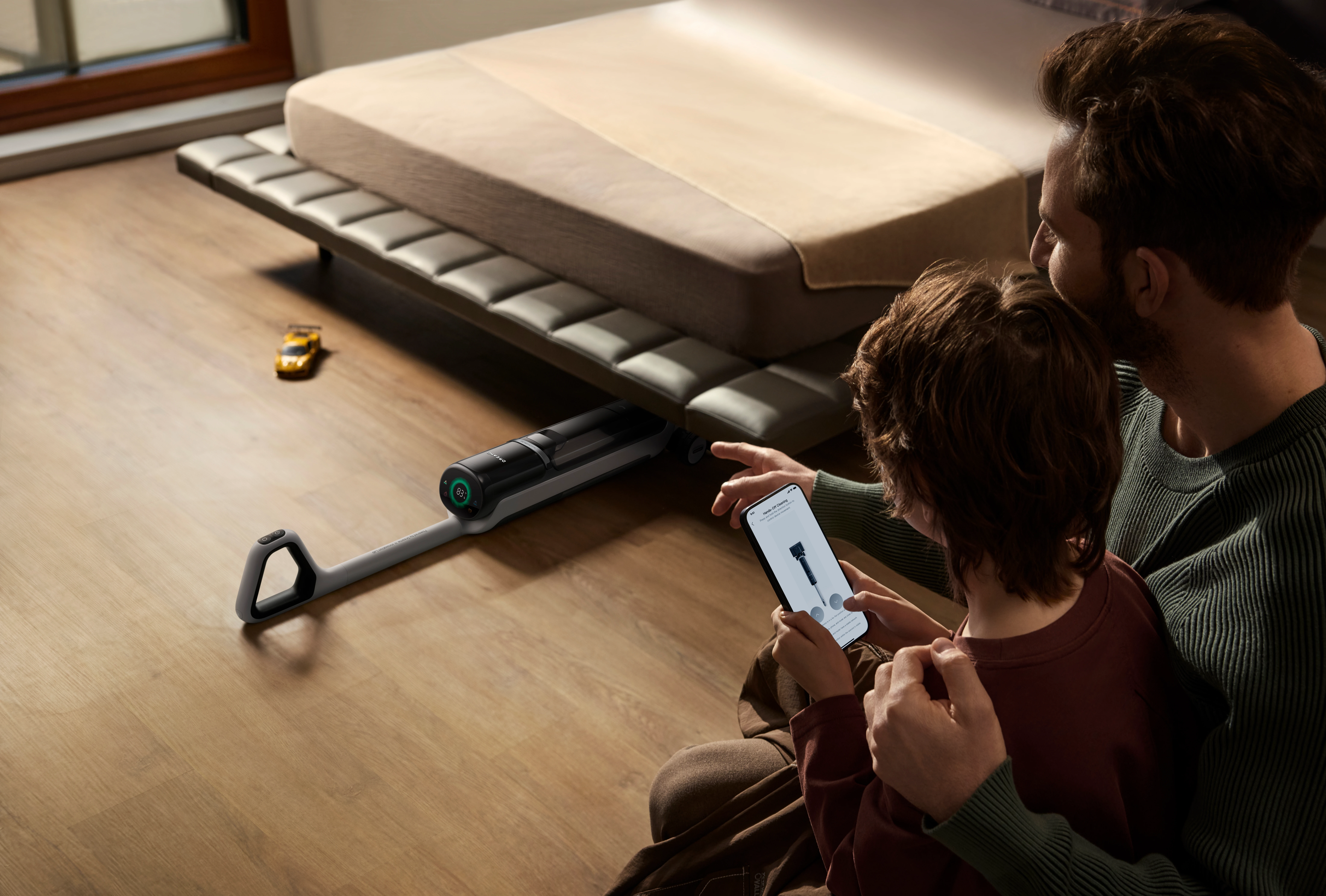 Mężczyzna i dziecko patrzą na smartfona obok łóżka. Odkurzacz leży na podłodze.