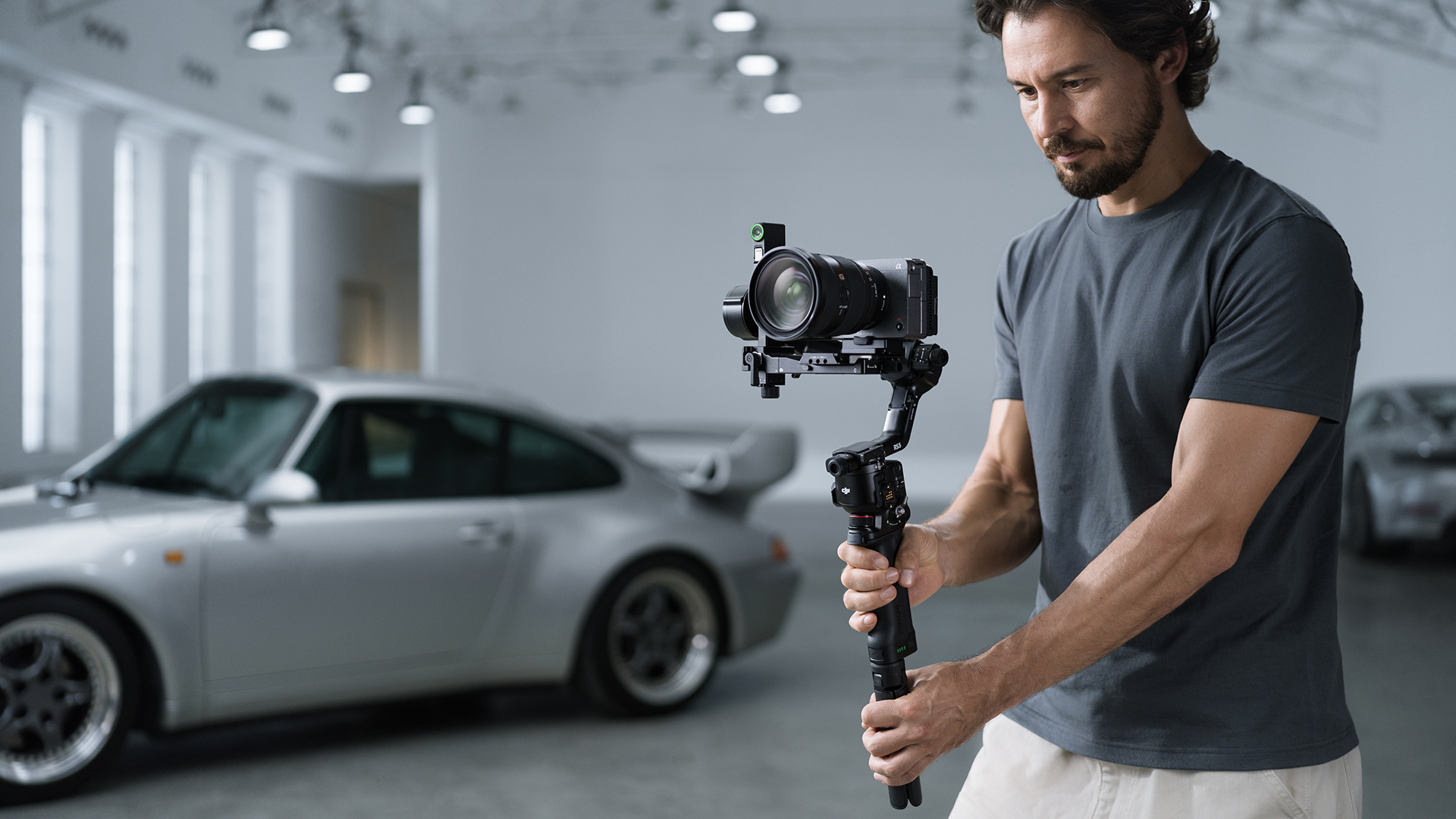 Adam parlak salonda gümüş spor arabanın önünde kamerayla gimbal tutuyor.