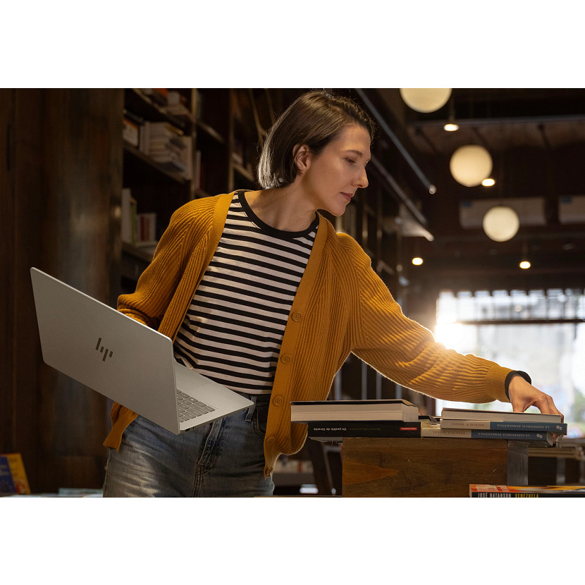 Kobieta z laptopem i żółtym kardiganem układa książki na półce.