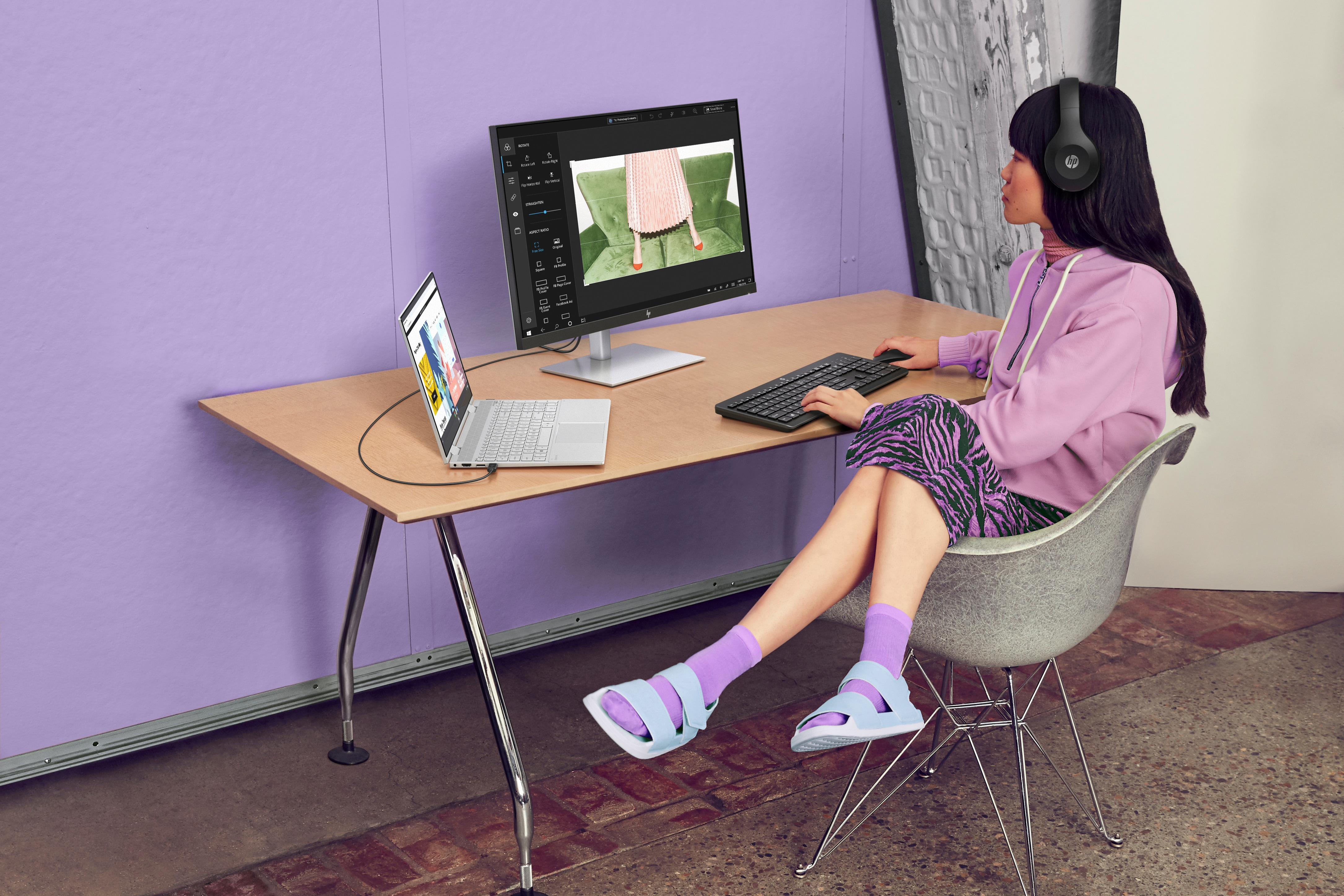 Kobieta w słuchawkach siedzi przy biurku z laptopem i monitorem.