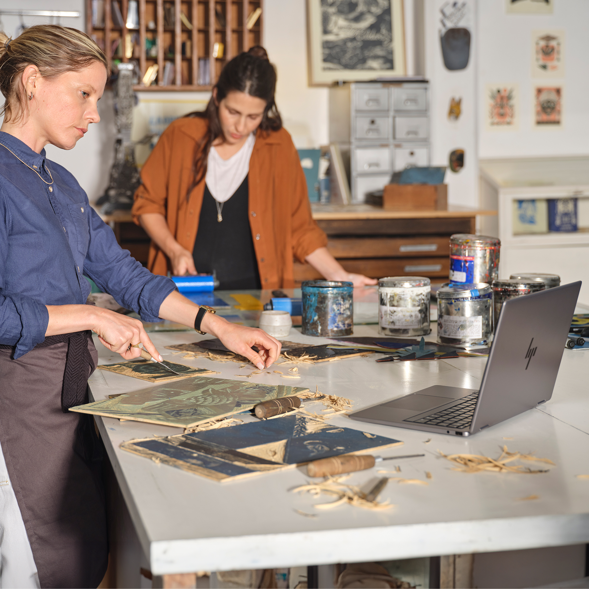 Dwie kobiety rzeźbią płyty drukarskie w pracowni z narzędziami i słoikami z farbą.