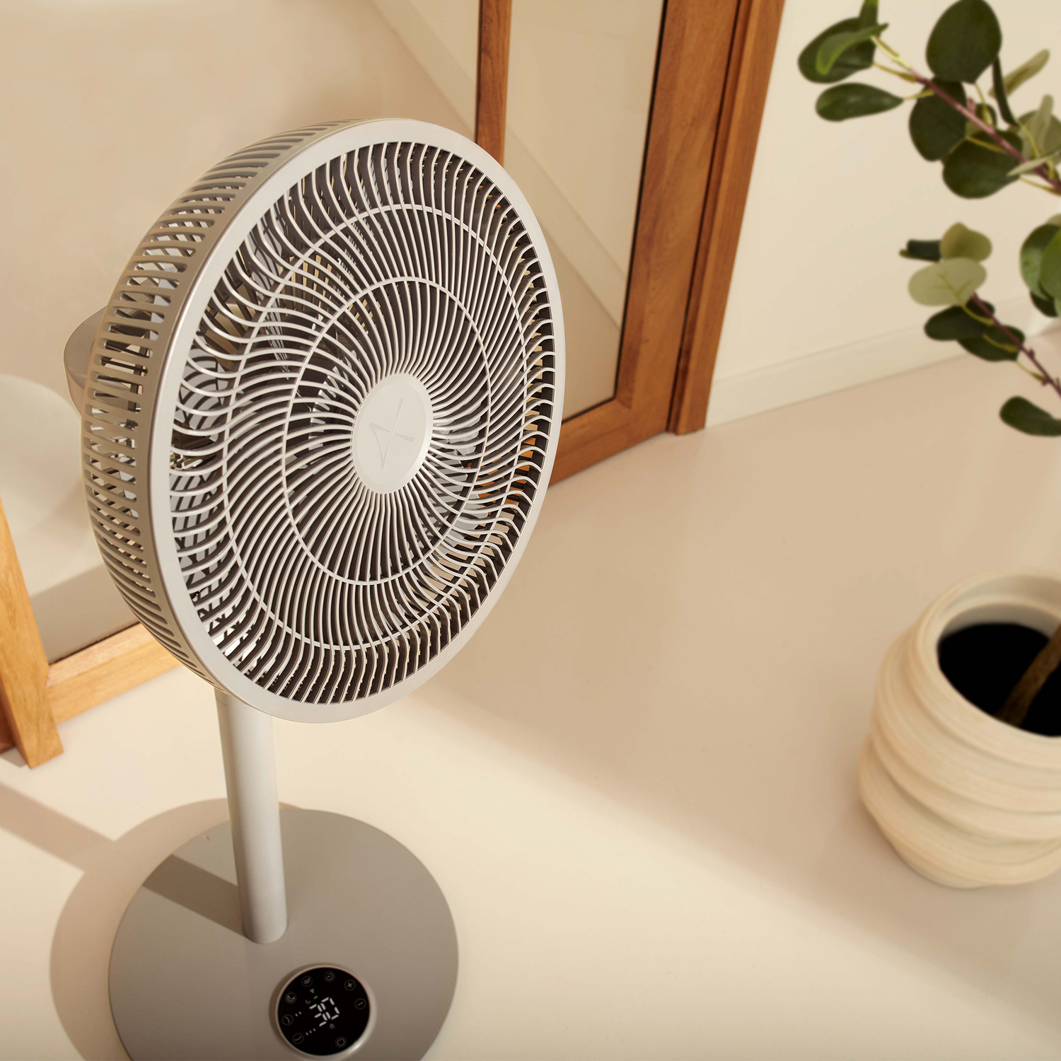 A gray standing fan with a digital display sits on a light floor next to a mirror and a plant.