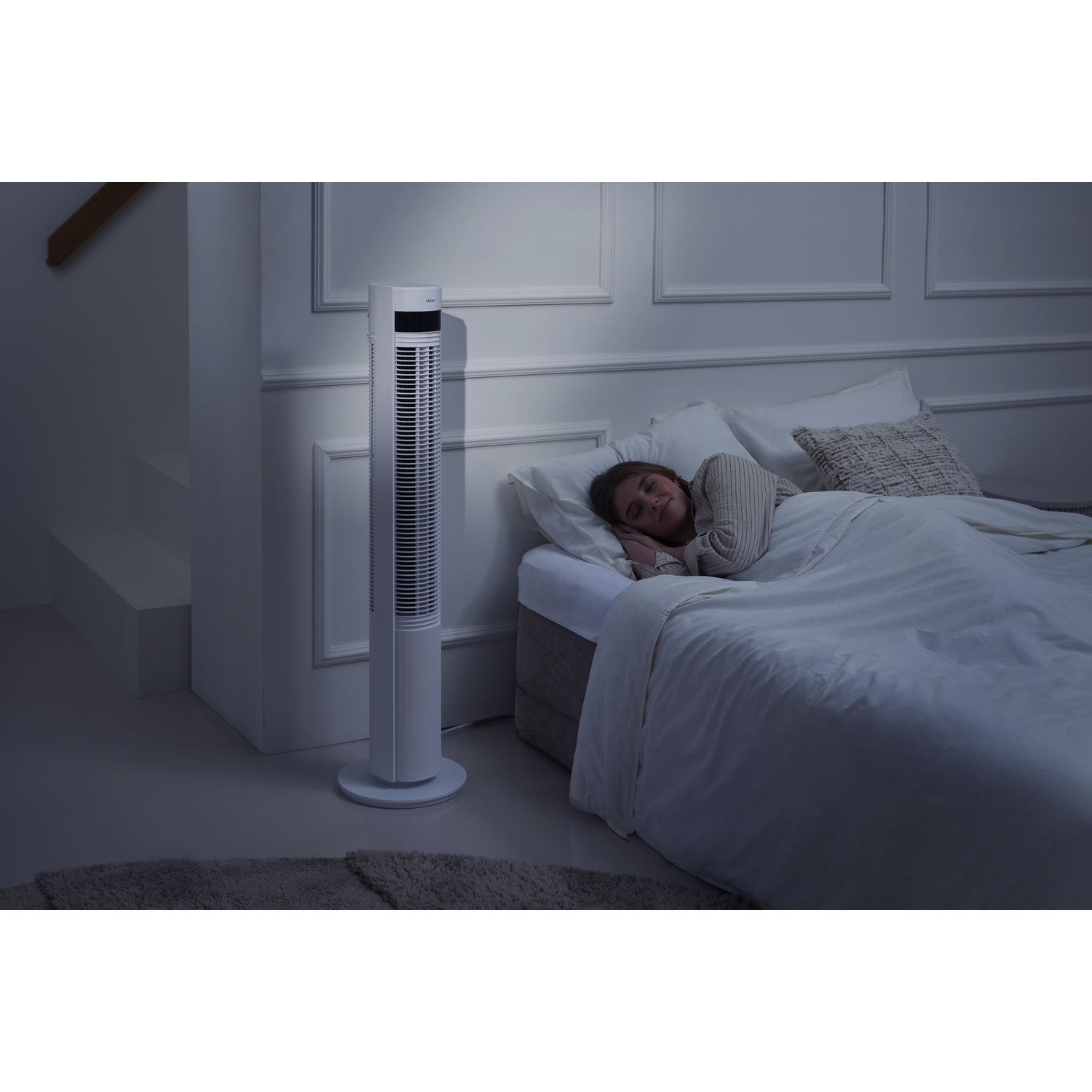 A woman sleeps in bed, a tower fan stands beside her.