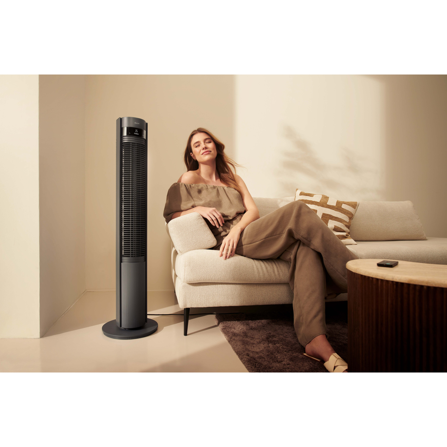 A woman sits on a sofa next to a slim tower fan. A coffee table is nearby.