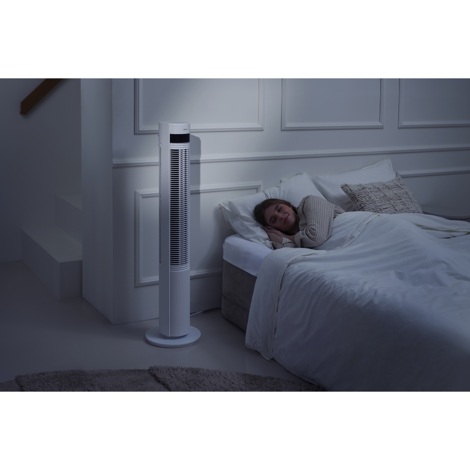 A woman sleeps in bed while a white tower fan operates in a dark room.