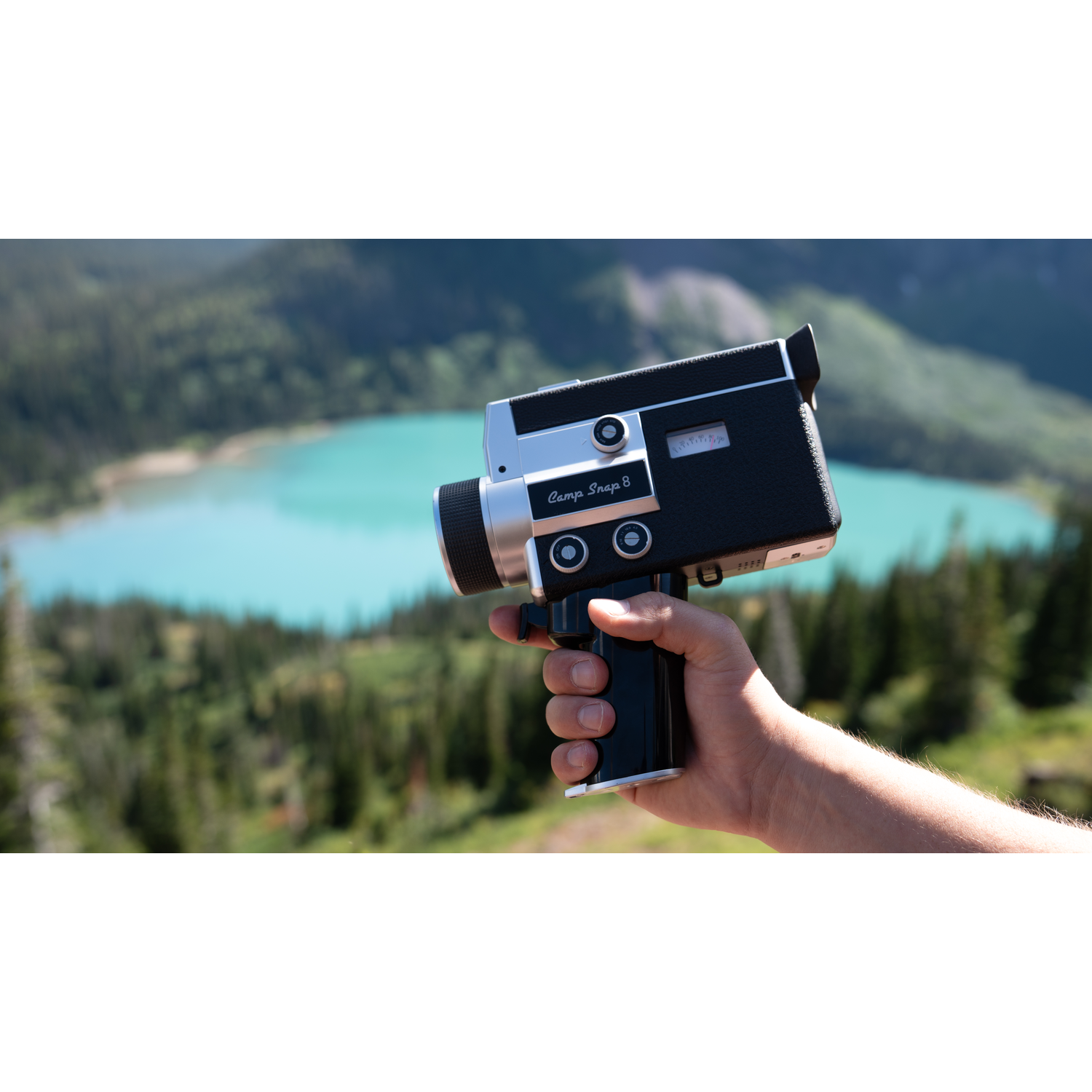 Een hand houdt een vintage camera vast voor een turkooizen meer en beboste bergen.