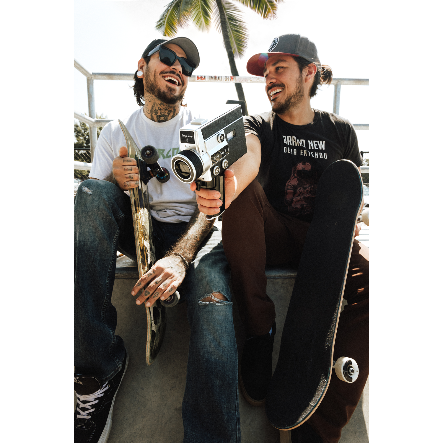 Twee lachende mannen zitten met skateboards en een camera.