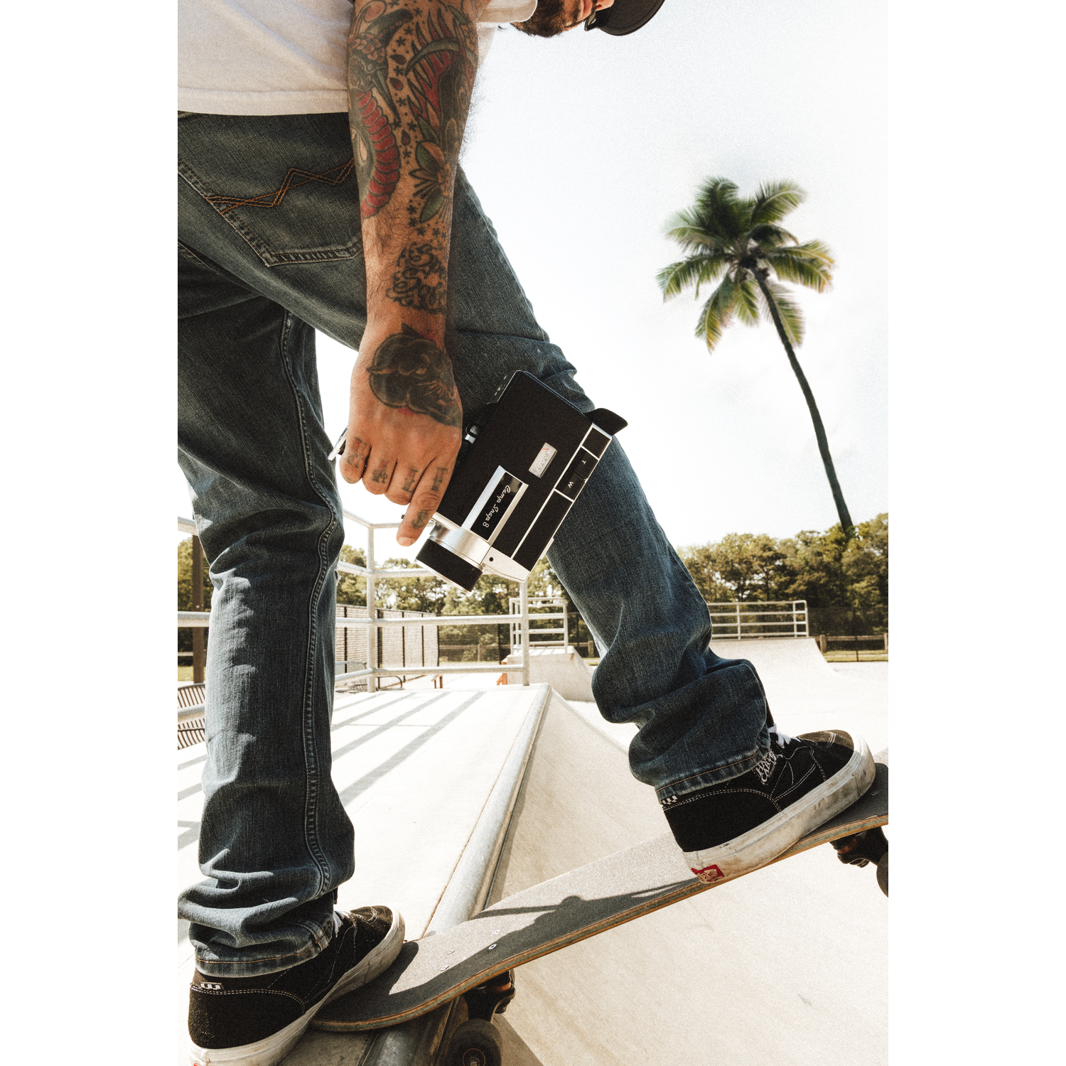 Een man met getatoeëerde armen houdt een camera vast op een skateboard in een skatepark.
