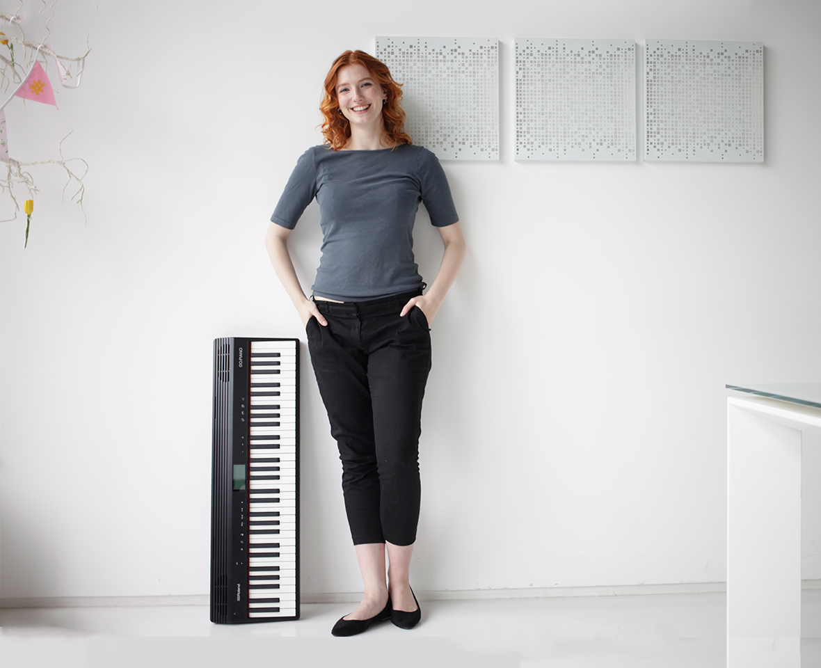 Une rousse se tient à côté d'un clavier noir devant un mur blanc.
