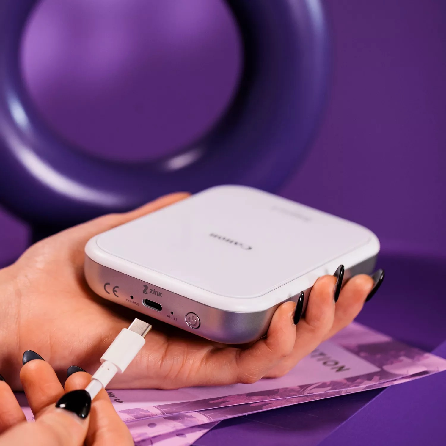 Dłoń z czarnymi paznokciami podłącza biały kabel USB do białej drukarki.